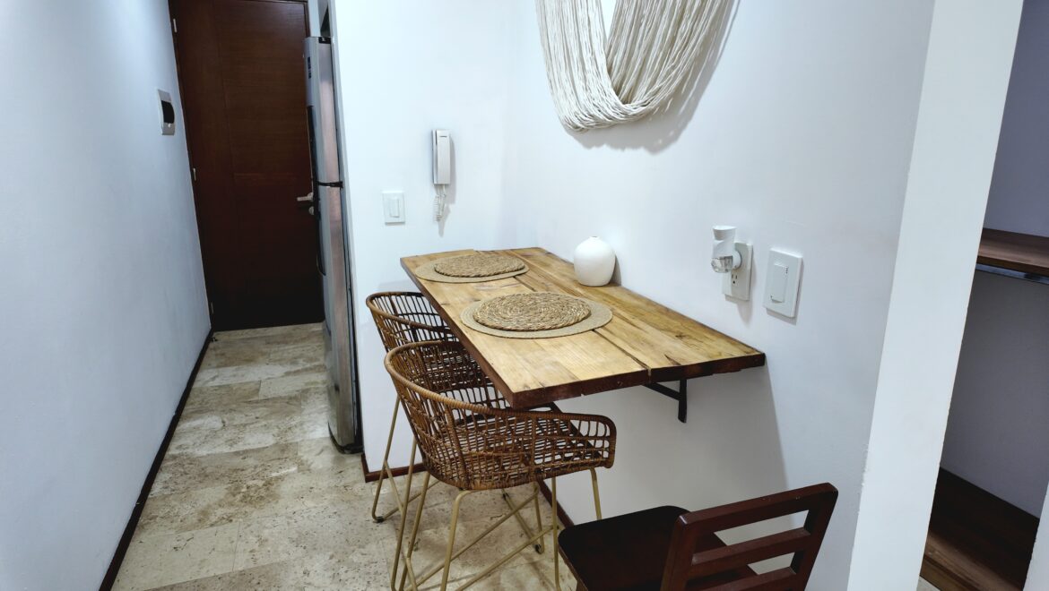 Narrow dining nook with a rustic wooden table, two wicker chairs, woven placemats, and a white vase against white walls; macrame wall hanging overhead.