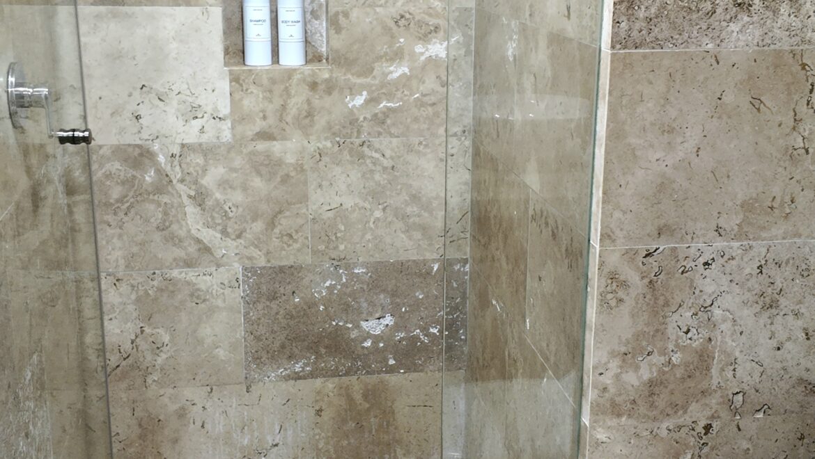 Glass-walled shower with beige marble tiles, built-in niche containing two white pump bottles, and a white toilet nearby in a modern bathroom.