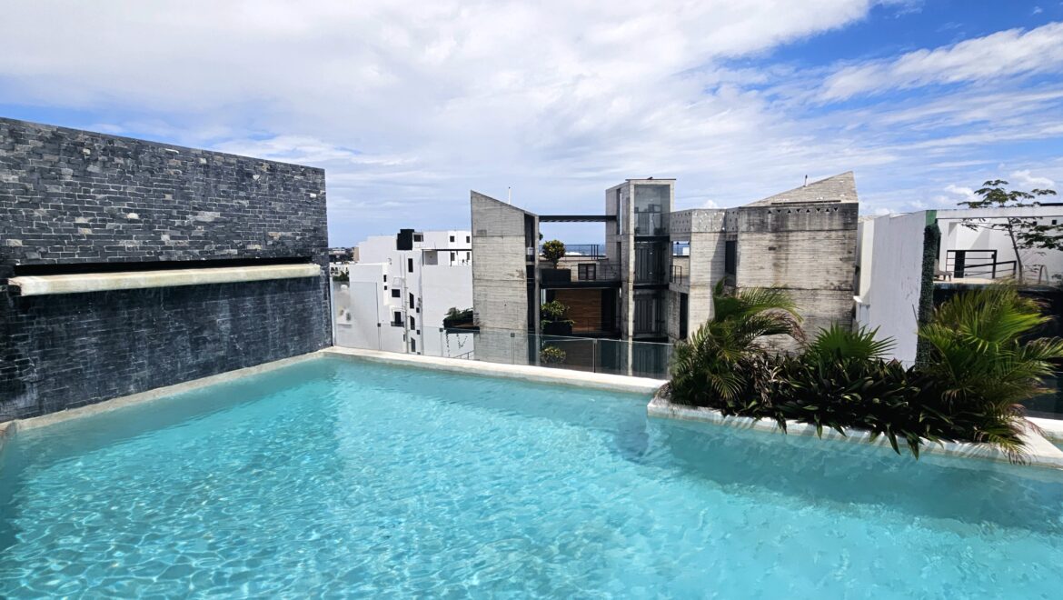 Rooftop swimming pool with turquoise water, modern stone wall, and city buildings in the background beneath a blue sky.