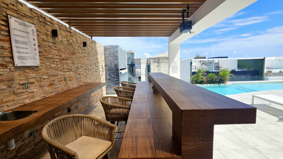 Outdoor poolside bar area with a long wooden counter, wicker chairs, and a stone wall under a wooden pergola, overlooking a bright blue pool and sky.