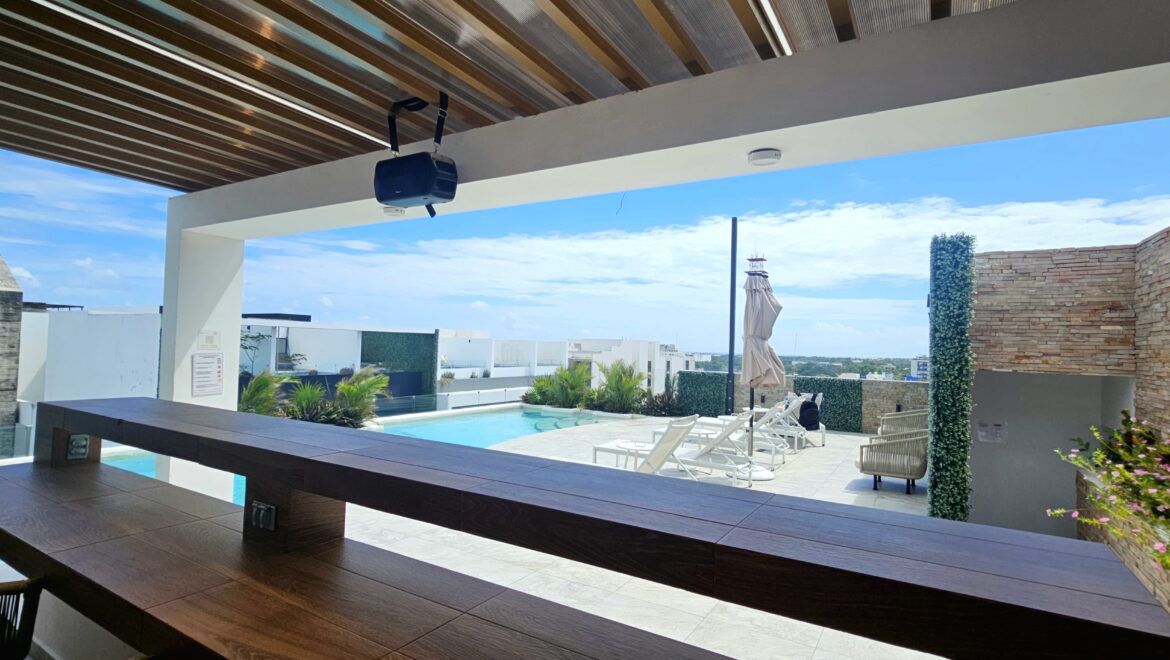 Rooftop pool area with white lounge chairs, an umbrella, and stone walls under a blue sky.