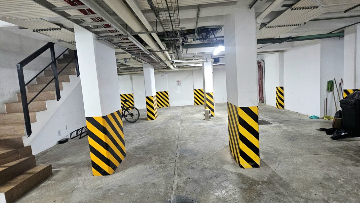 Underground parking level with concrete pillars painted white and black-yellow hazard stripes, stairs on the left, and scattered items.