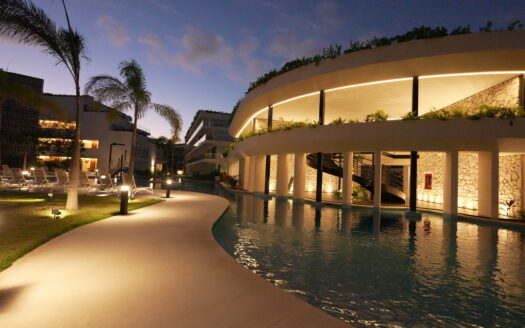 Resort pool area at dusk with modern curved building, palm trees, and illuminated walkway lights by the water.