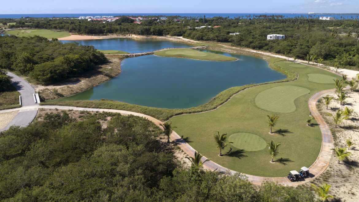 Aerial view of a golf course with winding cart paths, palm trees, and teal lagoons bordered by sandy areas and green fairways near a distant coastline.