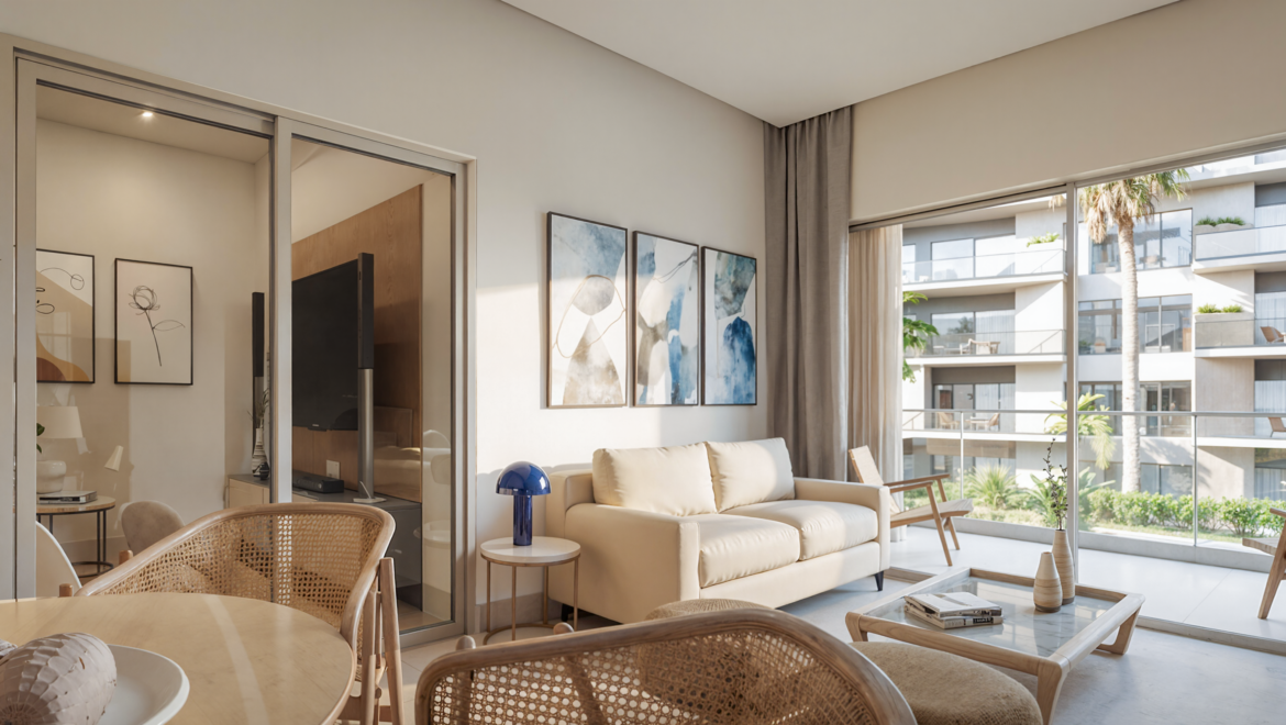 Bright living room with beige sofa, blue-accent wall art, and a glass balcony offering greenery and neighboring apartments outside.