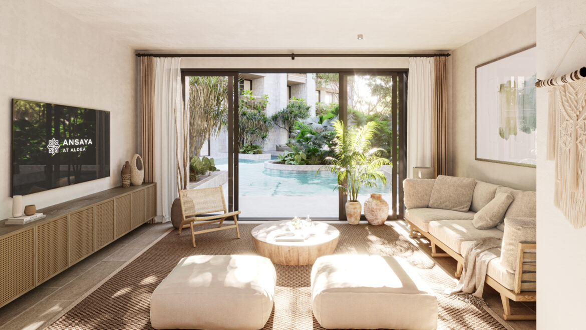 Living room with neutral furniture and a large sliding glass door opening to a pool and lush tropical garden outside. On the left is a wall-mounted TV showing the logo 'ANSAYA AT ALDEA'.