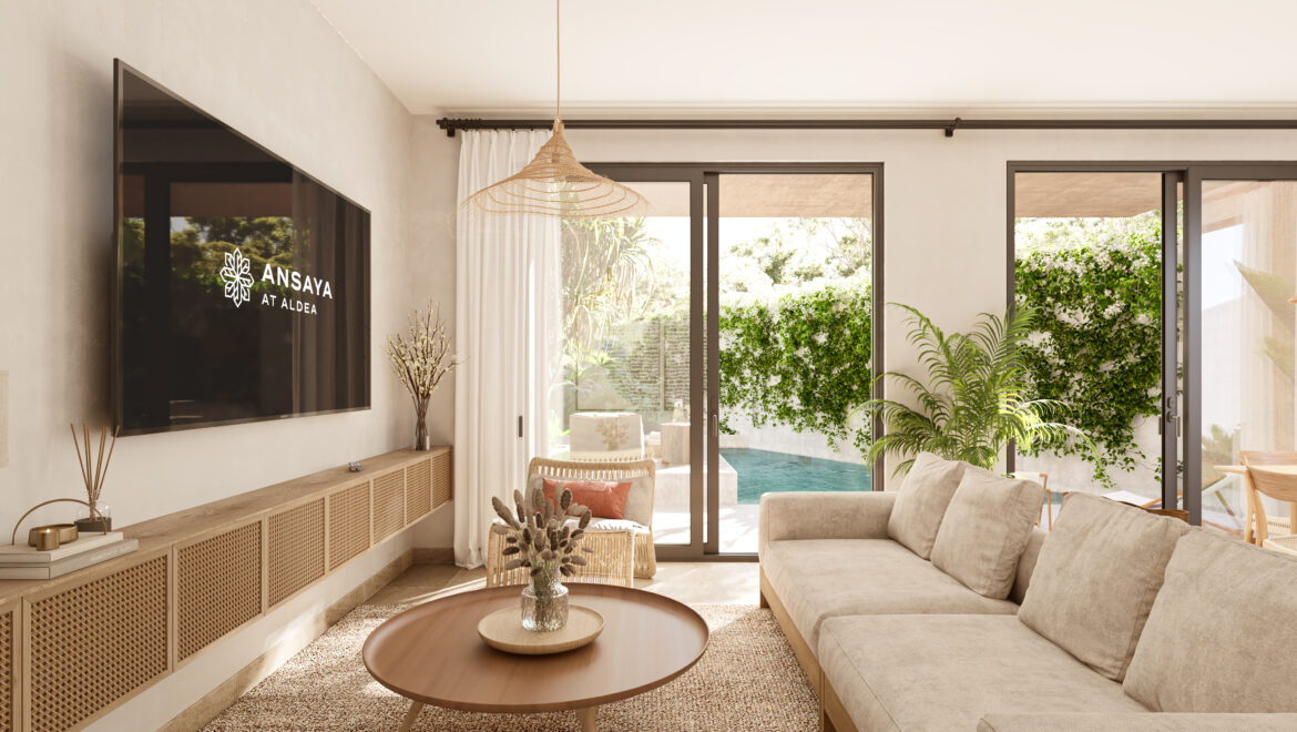 Living room with a beige sectional, round wooden coffee table, and wall-mounted TV showing 'ANSAVA AT ALDEA,' with glass doors opening to a sunny pool area.