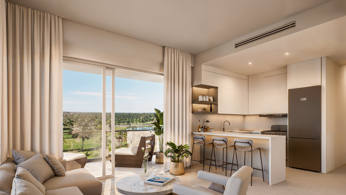 Open-plan living room and kitchen in a modern apartment, featuring a beige sectional sofa, round marble coffee table, and bar stools by a white island with a tropical view outside the sliding doors.