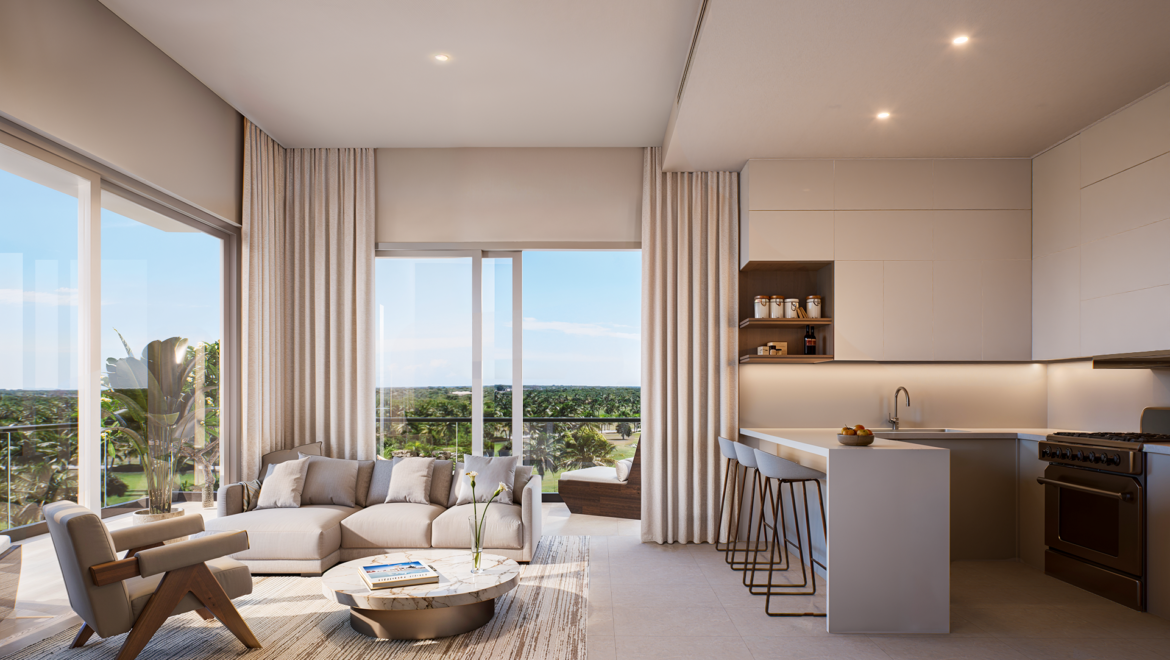 Open-plan living area with a beige sectional, round marble coffee table, and large windows overlooking a tropical landscape, connected to a modern kitchen island.