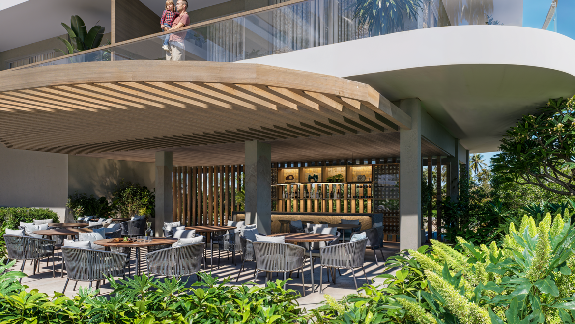 Open-air restaurant with wooden slatted ceiling, glass balcony above, and lush tropical plants surrounding the dining area; man holds child on the balcony railing.