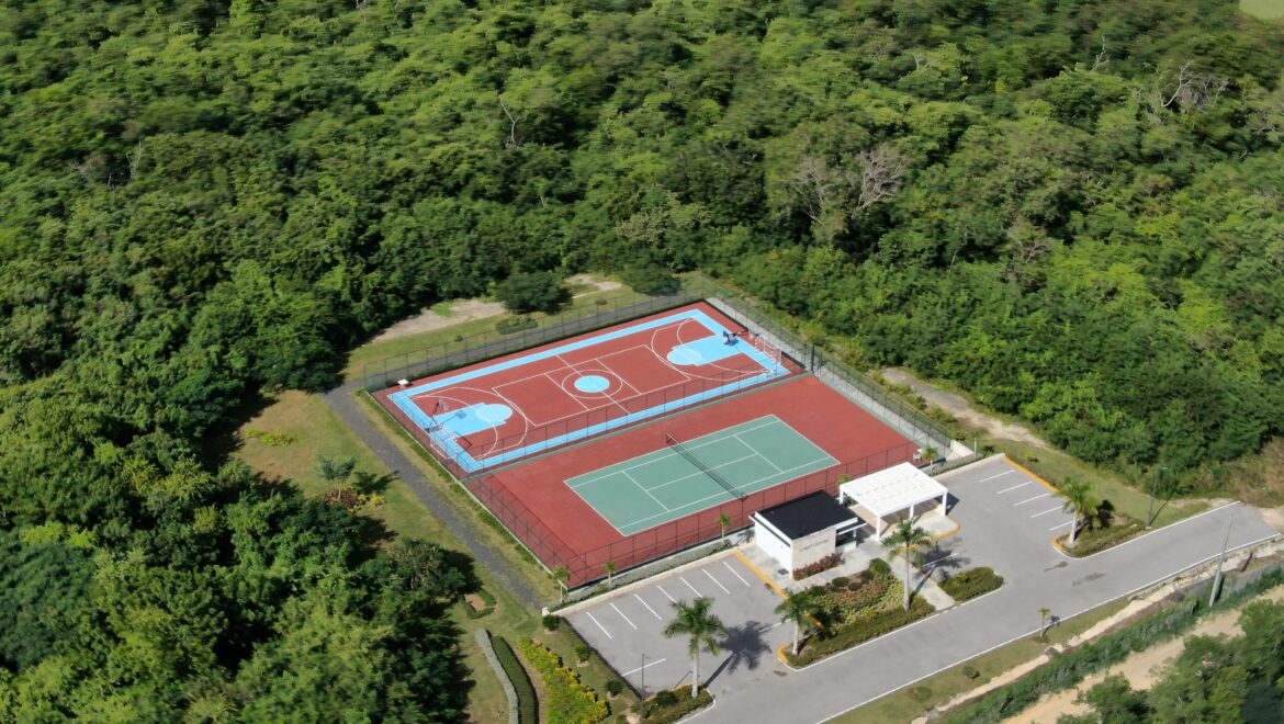 Aerial view of a fenced sports court complex with red surfaces and blue markings, surrounded by dense green trees and nearby roads.