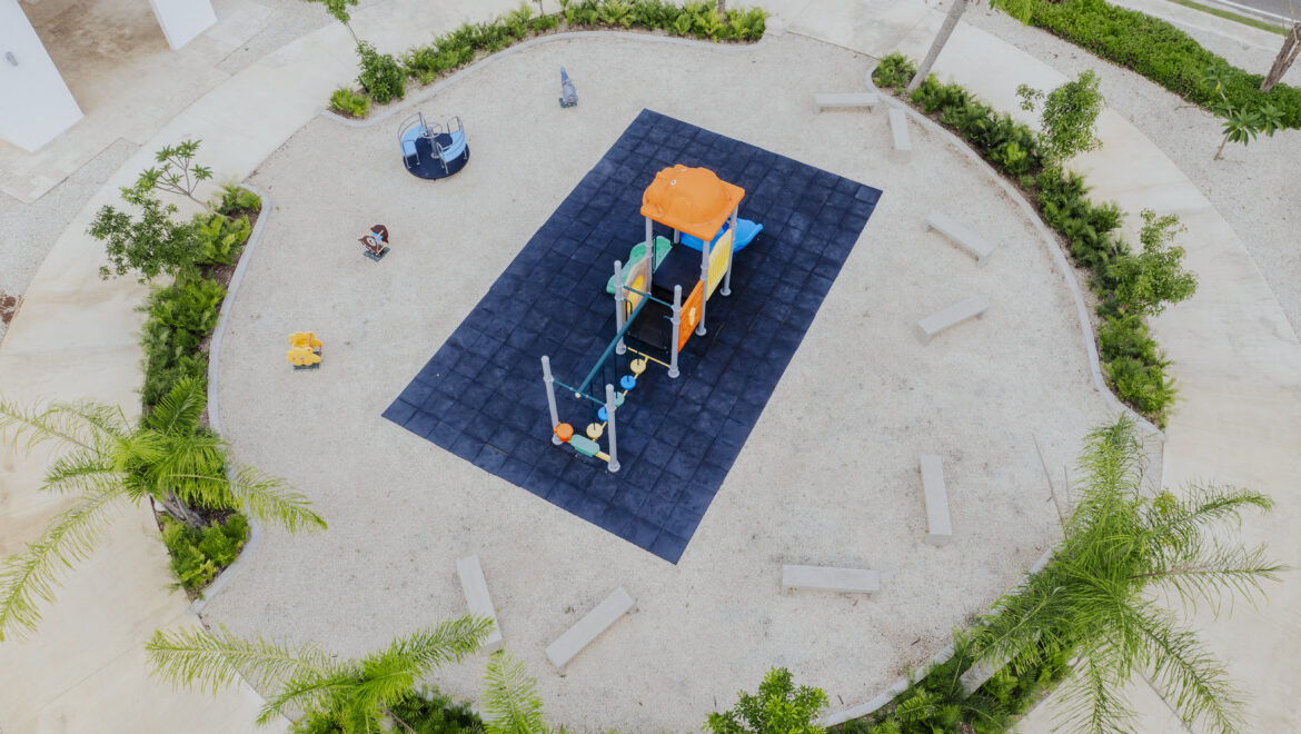 Aerial view of a circular park with a colorful playground on a dark blue surface, orange slide and climbing structure, surrounded by gravel and greenery.