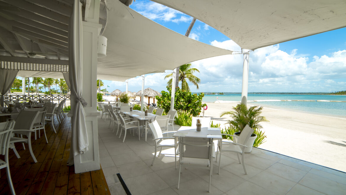 Outdoor beachside restaurant with white tables and chairs under white shade sails, palm trees and turquoise sea visible beyond.