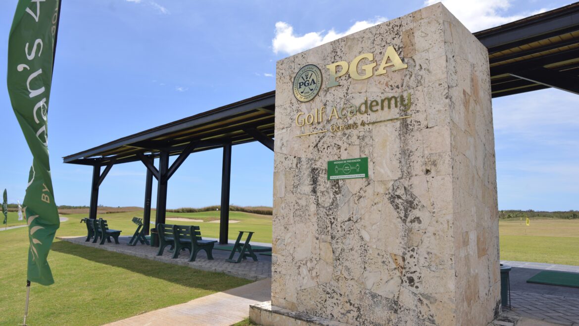 PGA Golf Academy sign on a stone pillar at a golf course with green flag and seating under a shaded structure.