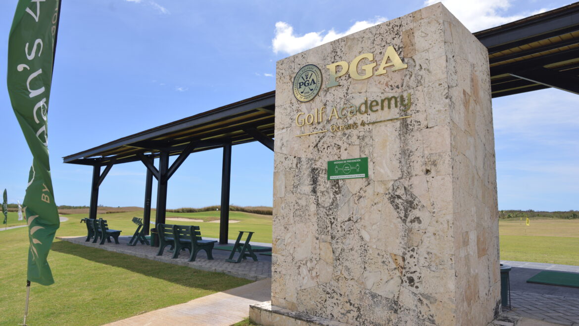 PGA Golf Academy sign on a stone pillar with a green flag beside a sunny practice area at a golf course