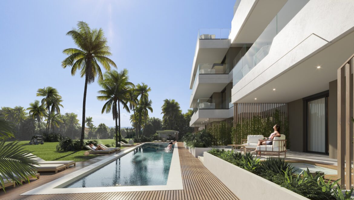 Modern white apartment building with glass balconies, poolside deck, and tall palm trees under a bright blue sky.