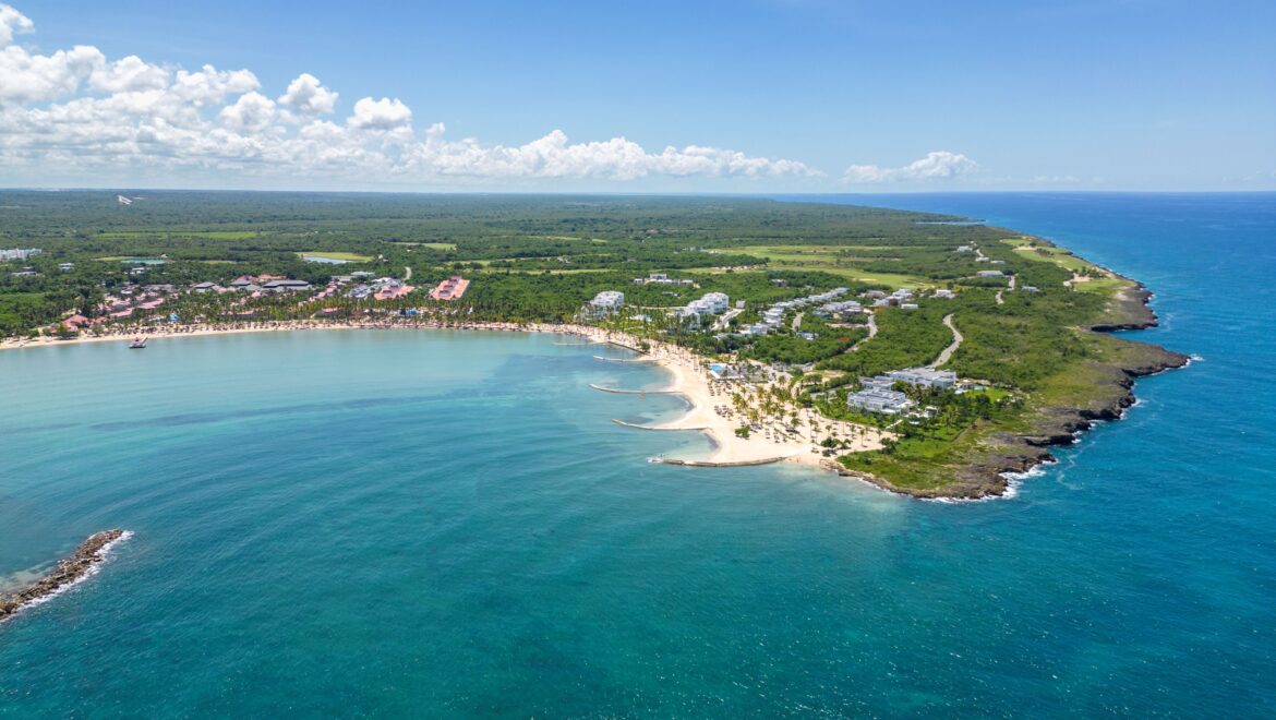 Aerial view of a tropical coastline with a curved white-sand beach, palm trees, and turquoise sea meeting green land far inland.
