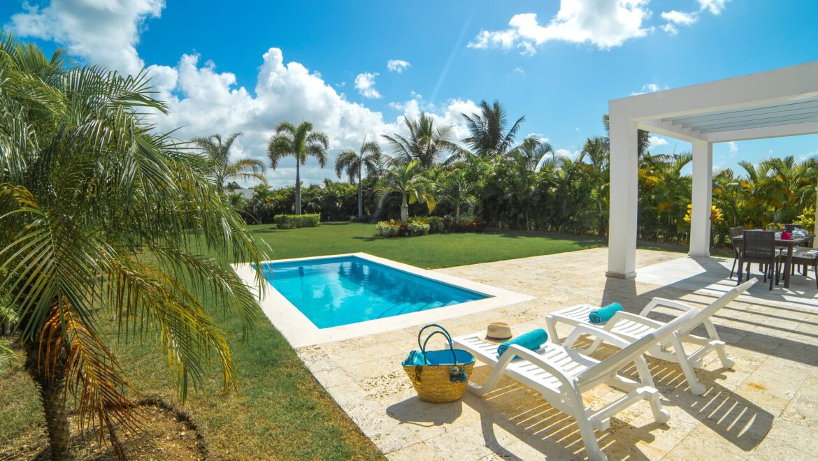 Sunny backyard pool scene with blue water, white stone surround, and lounge chairs under a bright blue sky with palm trees nearby.