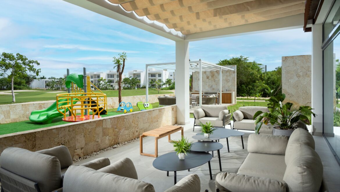 Outdoor patio with beige cushioned seating under a pergola; modern glass walls and stone accents, sunny day.