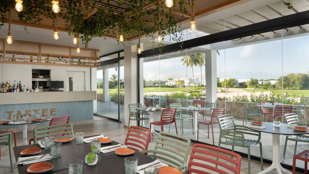 Bright restaurant interior with hanging Edison bulbs, trailing greenery, and colorful metal chairs around dark tables set for service, glass walls view outdoors to palm trees.