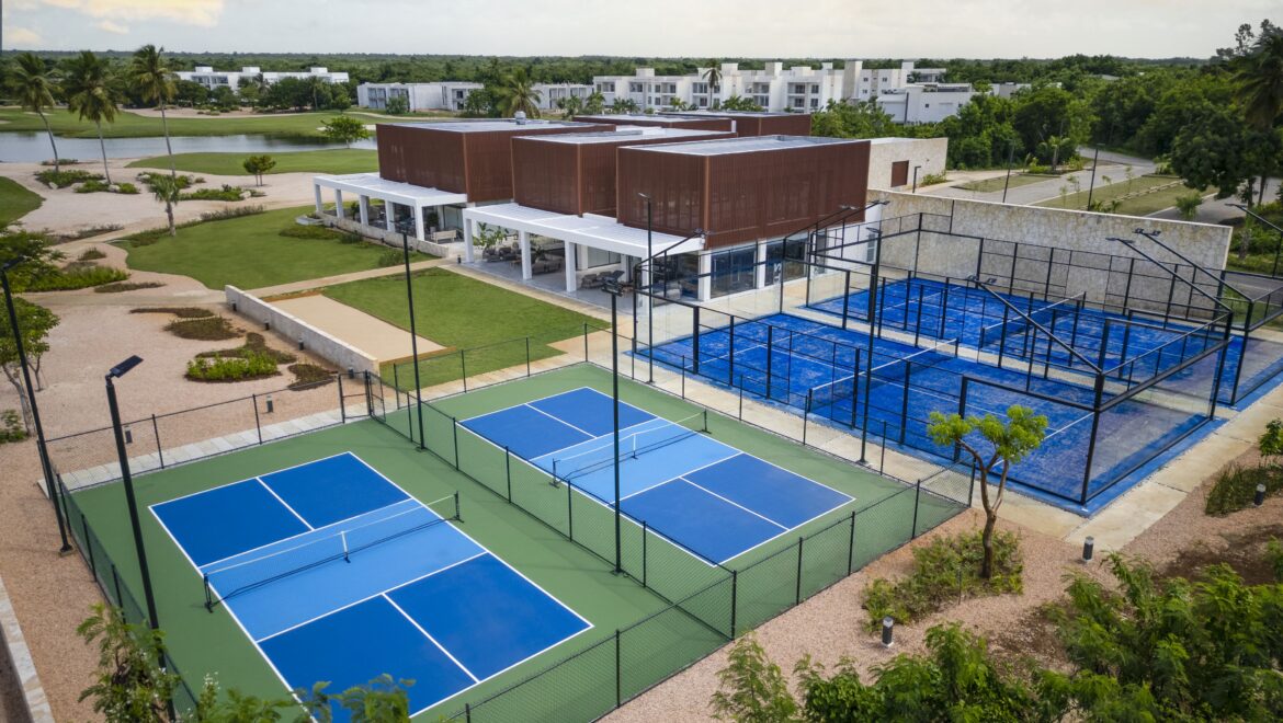 Aerial view of a sports complex with blue tennis courts, green lawns, and a brown modern building surrounded by trees.