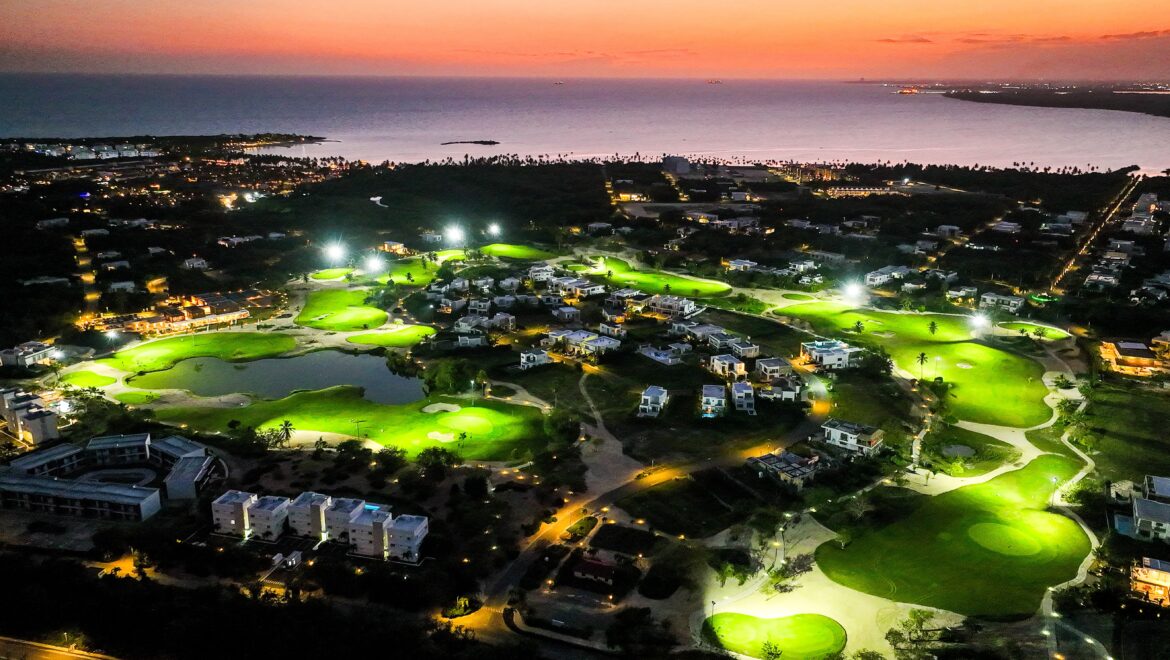 Aerial view of a coastal town at sunset with a lit golf course in the foreground and calm sea on the horizon.