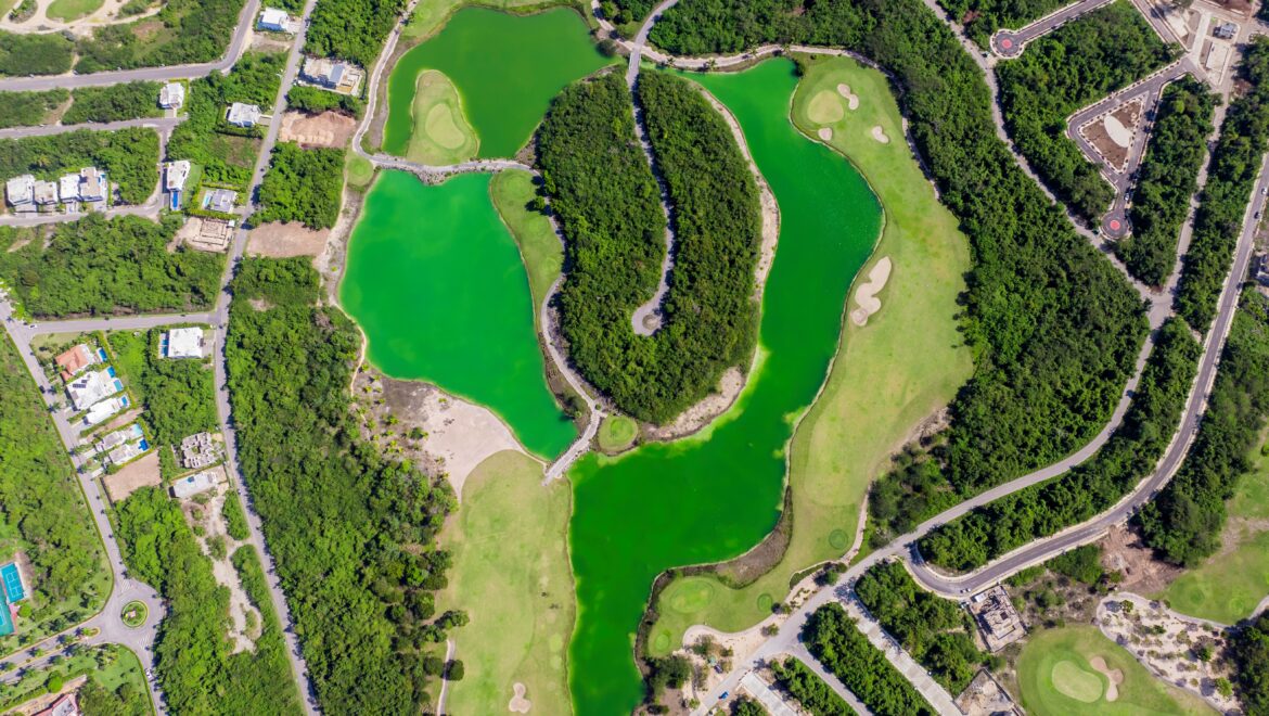 Aerial view of a winding golf course with green fairways, sand traps, and bright green water features bordered by trees and roads nearby.