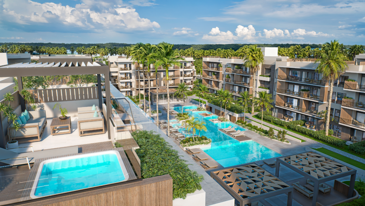 Aerial view of a modern resort with a large central pool, lounge chairs, and palm trees between beige apartment buildings.
