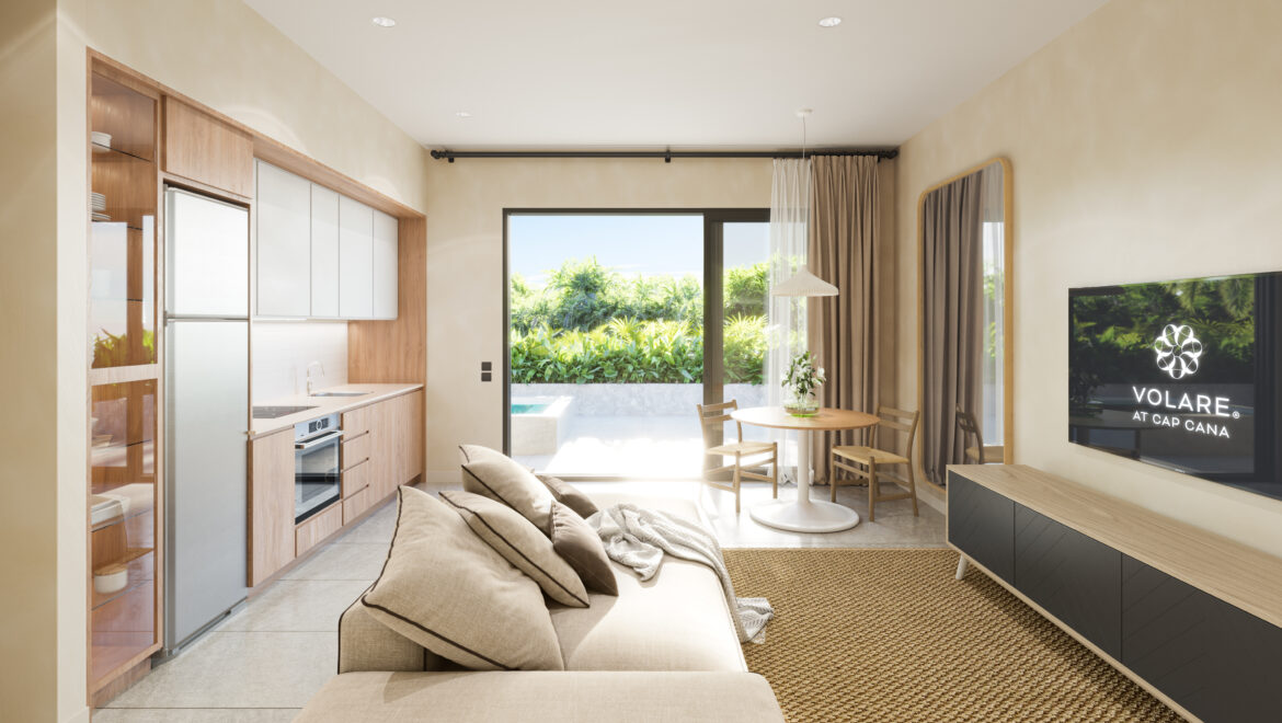 Bright open-plan living space with a kitchen on the left, beige sofa in the foreground, round dining table, and a wall-mounted TV reading 'VOLARE AT CAP CANA'.