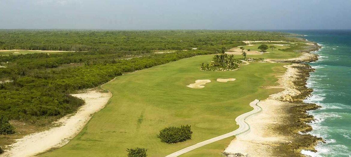 Aerial view of a coastal golf course with green fairways along a rocky shoreline and turquoise sea to the right.