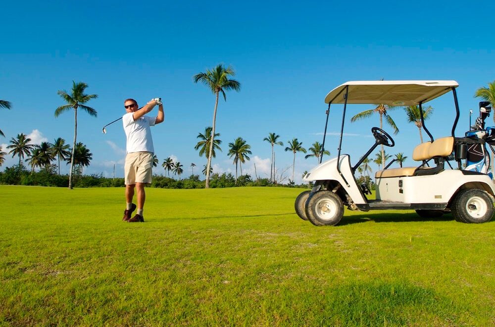 A man in a white shirt and khaki shorts swings a golf club on a sunny golf course beside a white golf cart.