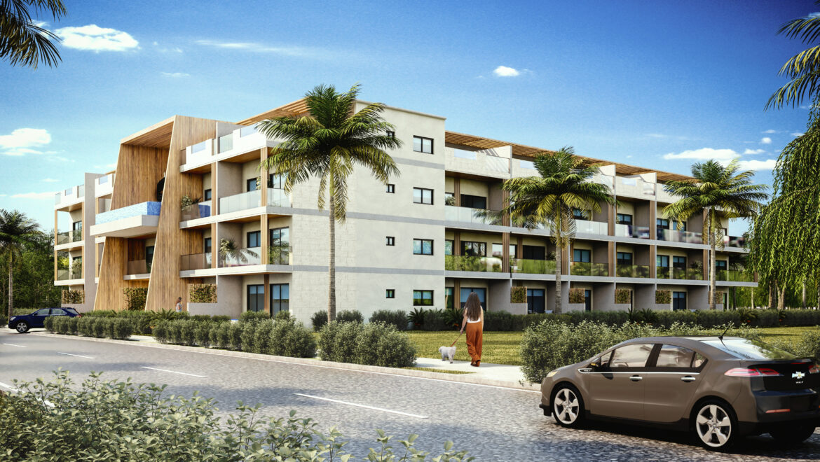 Modern four-story apartment building with balconies and palm trees along a cobblestone street, under a clear blue sky.