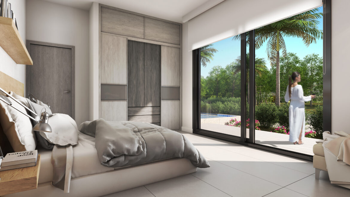 Woman in a white robe stands by floor-to-ceiling glass doors looking outside to a tropical garden with palm trees, bedroom interior in view