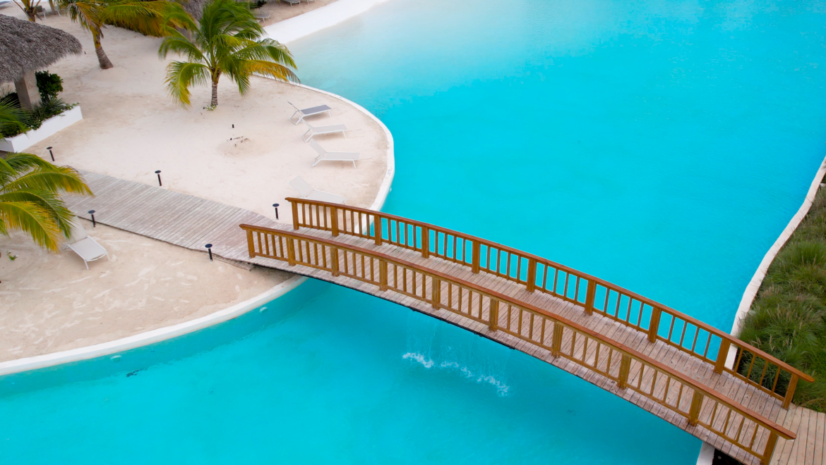 Aerial view of a tropical beach with white sand, palm trees, and a curved wooden bridge over bright turquoise water.