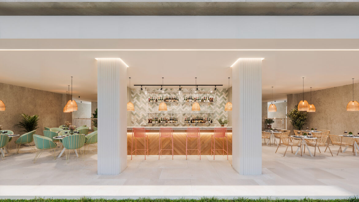 Modern, open-air bar with a central wine shelf, warm pendant lights, and pastel chairs on either side of a white counter.