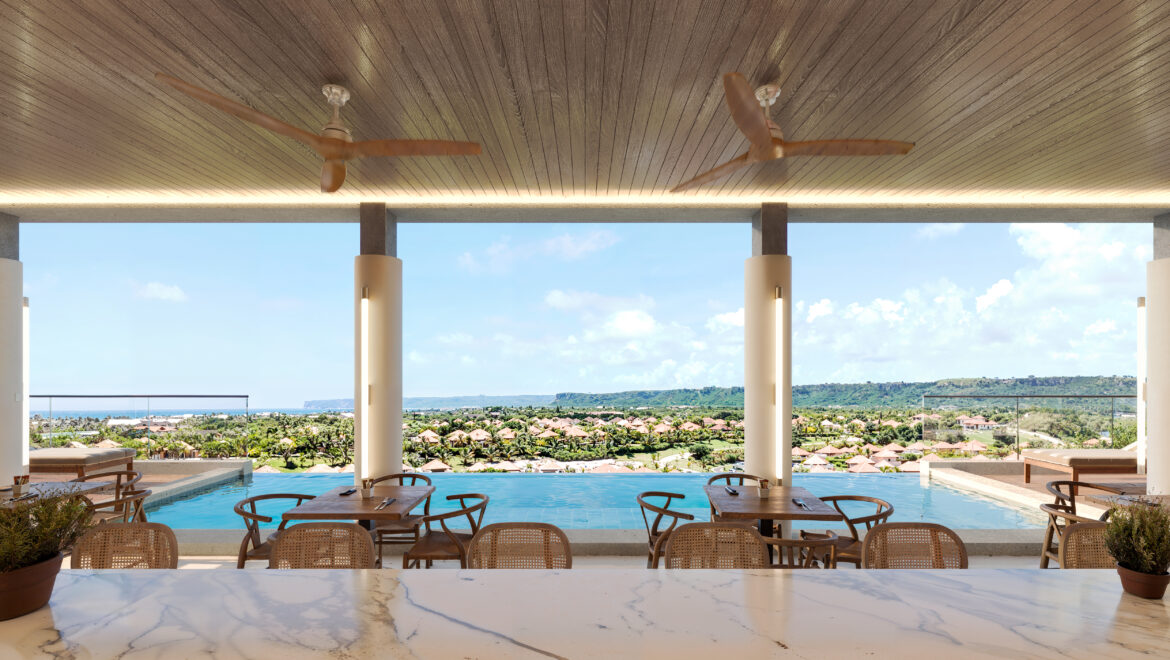 Open-air resort dining area with wooden tables and wicker chairs, a marble counter in the foreground, and ceiling fans, overlooking a pool and coastal landscape.