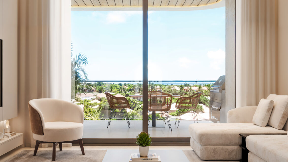 Living room with cream furniture opening to a balcony, featuring outdoor chairs and a view of palm trees and the ocean beyond.