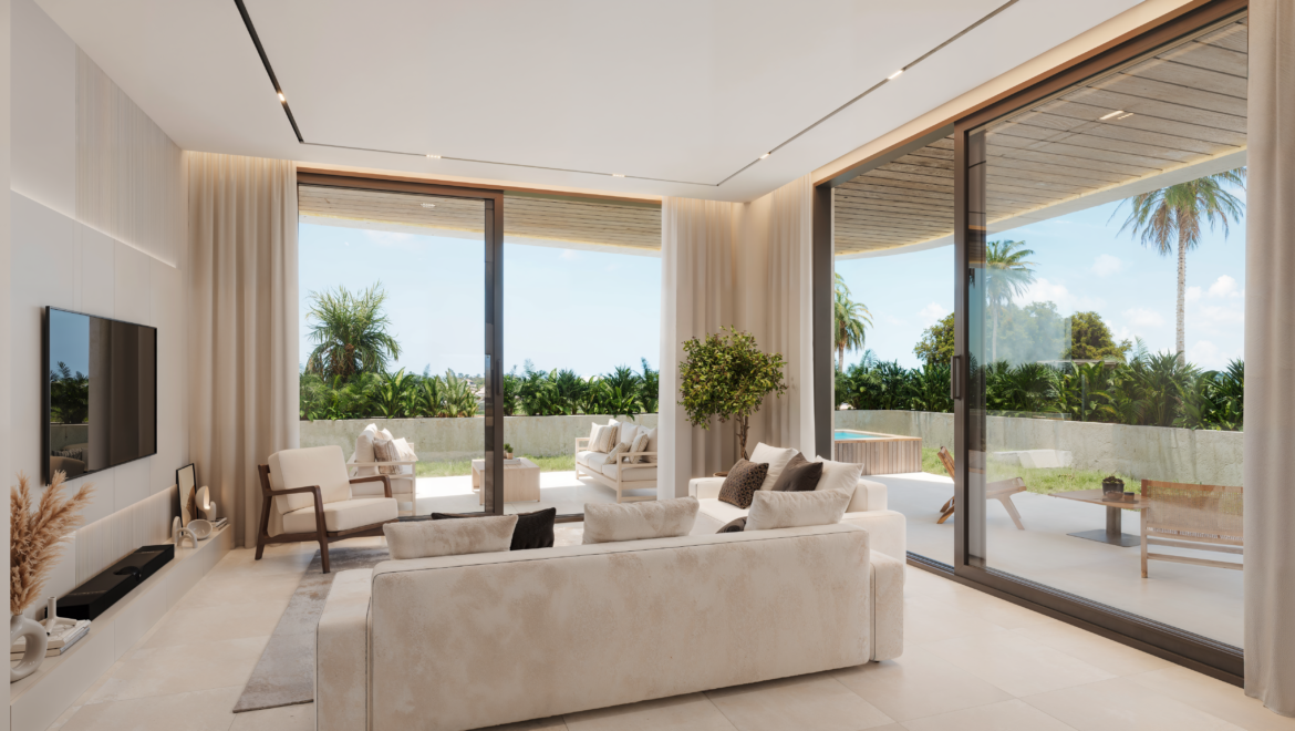 Bright living room with beige sofas, a wall TV, and floor‑to‑ceiling sliding glass doors opening to a tropical garden.