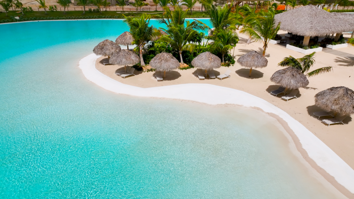 Aerial view of a tropical beach with turquoise water, white sand, and thatched umbrellas among palm trees.